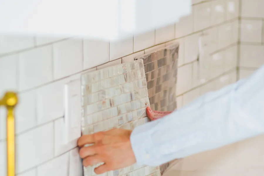 Mano presentando una pieza de mosaico cerámico frente a un muro revestido con azulejos.